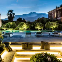 Un incantevole wine resort in Sicilia, con piscina illuminata, lettini e una vista mozzafiato sul vulcano al tramonto.