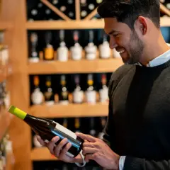 Uomo sorridente sceglie una bottiglia di vino, con scaffali pieni di bottiglie sullo sfondo. La sua unghia del vino è pronta per la degustazione.