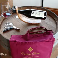 Bicchiere di vino rosso, cavatappi e borsa bordeaux con logo "Cascina Saria" su una botte di legno.