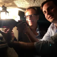 Two men in a wine cellar examine a bottle of wine, with a lantern in the background.
