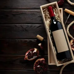 Scatola di legno con bottiglia di vino rosso, melograni e cavatappi, evocando i vini tipici della Campania.