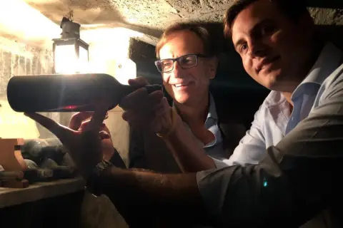 Two men in a wine cellar examine a bottle of wine, with a lantern in the background.