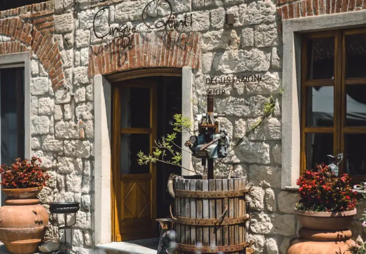 Ingresso di "Cinque Quinti", un negozio con insegna in ferro battuto, un torchio antico e vasi di fiori rossi.