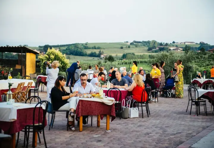 Un conviviale pranzo in vigna Monferrato, con tavoli apparecchiati all'aperto e persone che chiacchierano e gustano il cibo, sullo sfondo dolci colline.