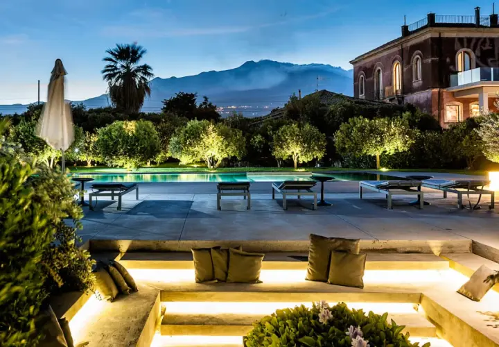 Un incantevole wine resort in Sicilia, con piscina illuminata, lettini e una vista mozzafiato sul vulcano al tramonto.