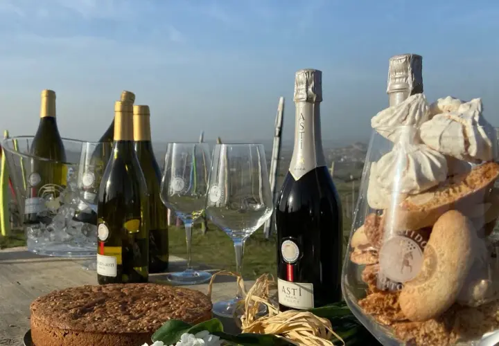 Bottiglie di vino dolce Asti, bicchieri, torta e dolcetti in un allestimento all'aperto.