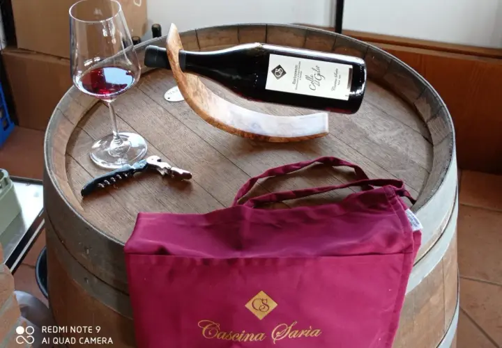 Bicchiere di vino rosso, cavatappi e borsa bordeaux con logo "Cascina Saria" su una botte di legno.