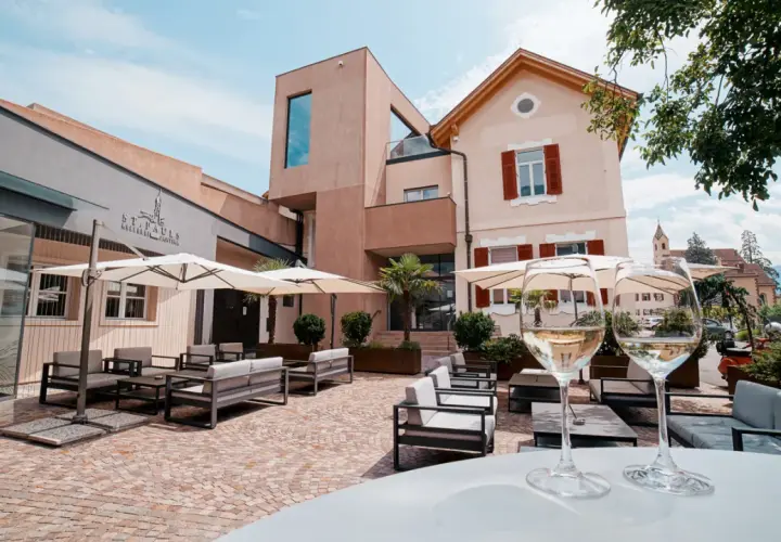 Terrazza accogliente con divani e ombrelloni, due calici di vino bianco in primo piano. La cantina St. Pauls sullo sfondo.