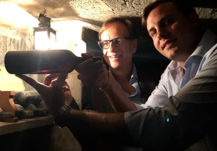 Two men in a wine cellar examine a bottle of wine, with a lantern in the background.