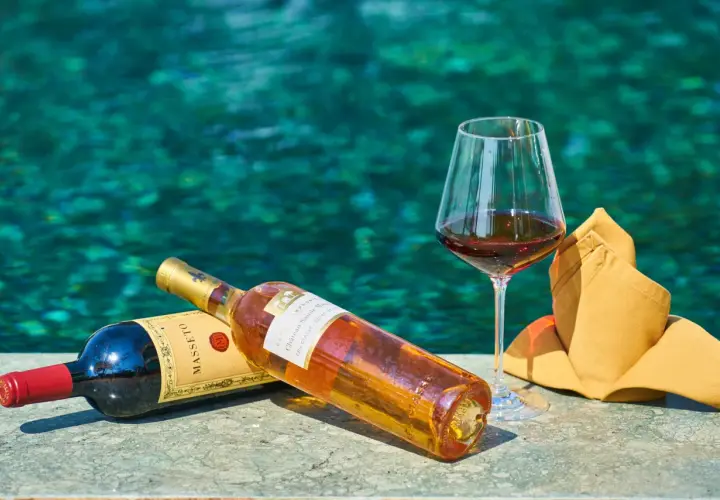 Due bottiglie di vino e un bicchiere pieno, con uno sfondo acquatico turchese. Un'immagine che evoca il sapido significato del relax e del piacere.