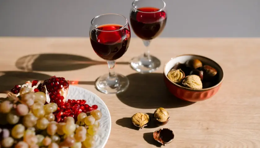 Due calici di vino rosso, uva, melograno e castagne, un'ambientazione che evoca un senso di abboccato significato, quasi un invito a gustare i piaceri semplici.