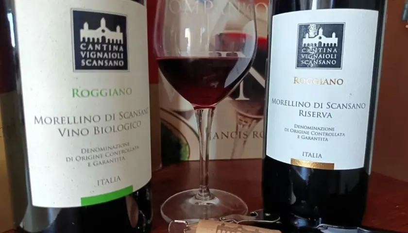 Due bottiglie di Morellino di Scansano e un calice di vino rosso, pronti per celebrare il santo del vino.