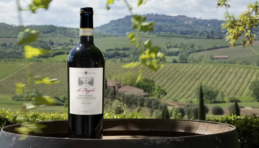 Bottiglia di Vino Nobile di Montepulciano Riserva su un barile, con vigneti e colline toscane sullo sfondo. Ideale per una degustazione vini Montepulciano.