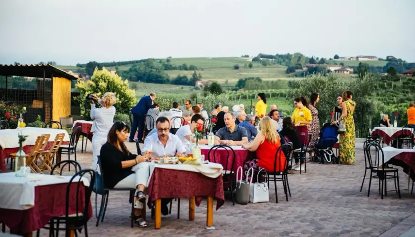 Un conviviale pranzo in vigna Monferrato, con tavoli apparecchiati all'aperto e persone che chiacchierano e gustano il cibo, sullo sfondo dolci colline.