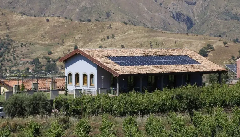 A light blue building with solar panels on its tiled roof sits behind a vineyard, with a dry, hilly landscape in the background.