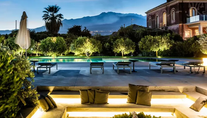 Un incantevole wine resort in Sicilia, con piscina illuminata, lettini e una vista mozzafiato sul vulcano al tramonto.