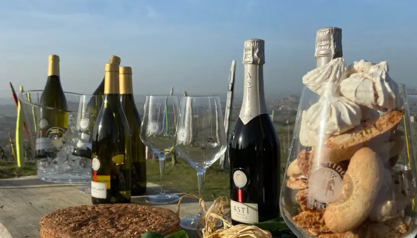 Bottiglie di vino dolce Asti, bicchieri, torta e dolcetti in un allestimento all'aperto.