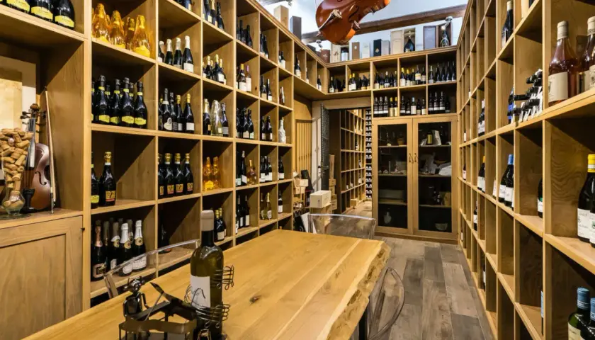 Cantine siciliane con scaffalature in legno piene di bottiglie di vino, un violino appeso e un tavolo in legno massiccio.