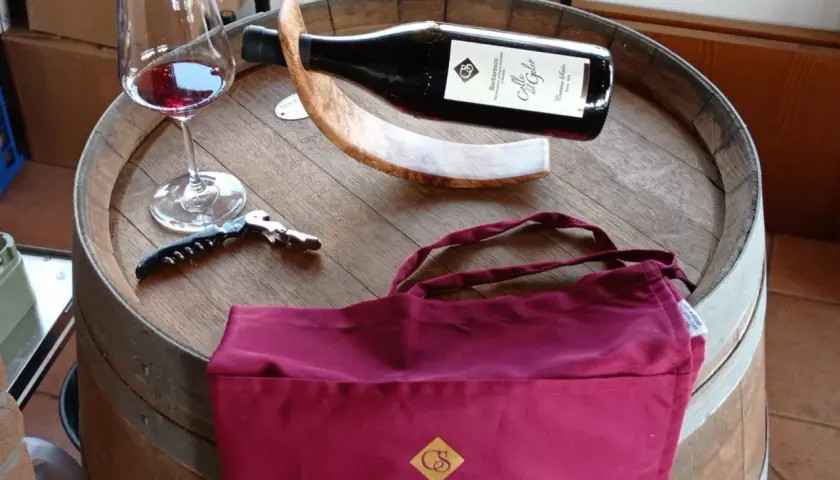 Bicchiere di vino rosso, cavatappi e borsa bordeaux con logo "Cascina Saria" su una botte di legno.