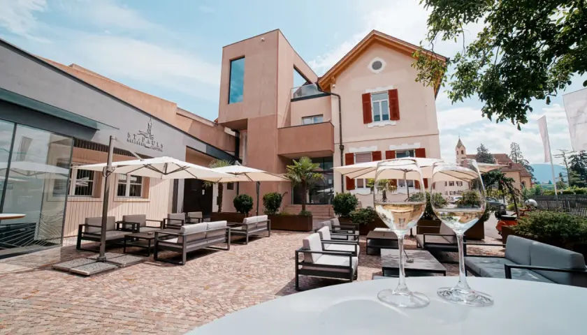Terrazza accogliente con divani e ombrelloni, due calici di vino bianco in primo piano. La cantina St. Pauls sullo sfondo.