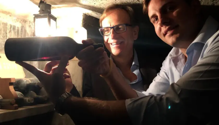 Two men in a wine cellar examine a bottle of wine, with a lantern in the background.