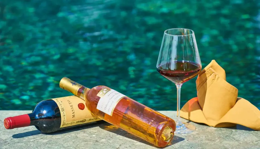 Due bottiglie di vino e un bicchiere pieno, con uno sfondo acquatico turchese. Un'immagine che evoca il sapido significato del relax e del piacere.