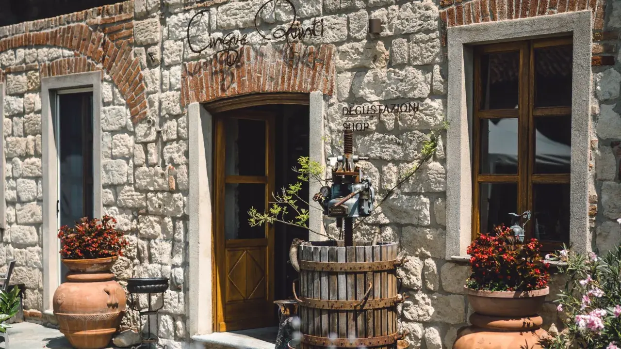 Ingresso di "Cinque Quinti", un negozio con insegna in ferro battuto, un torchio antico e vasi di fiori rossi.