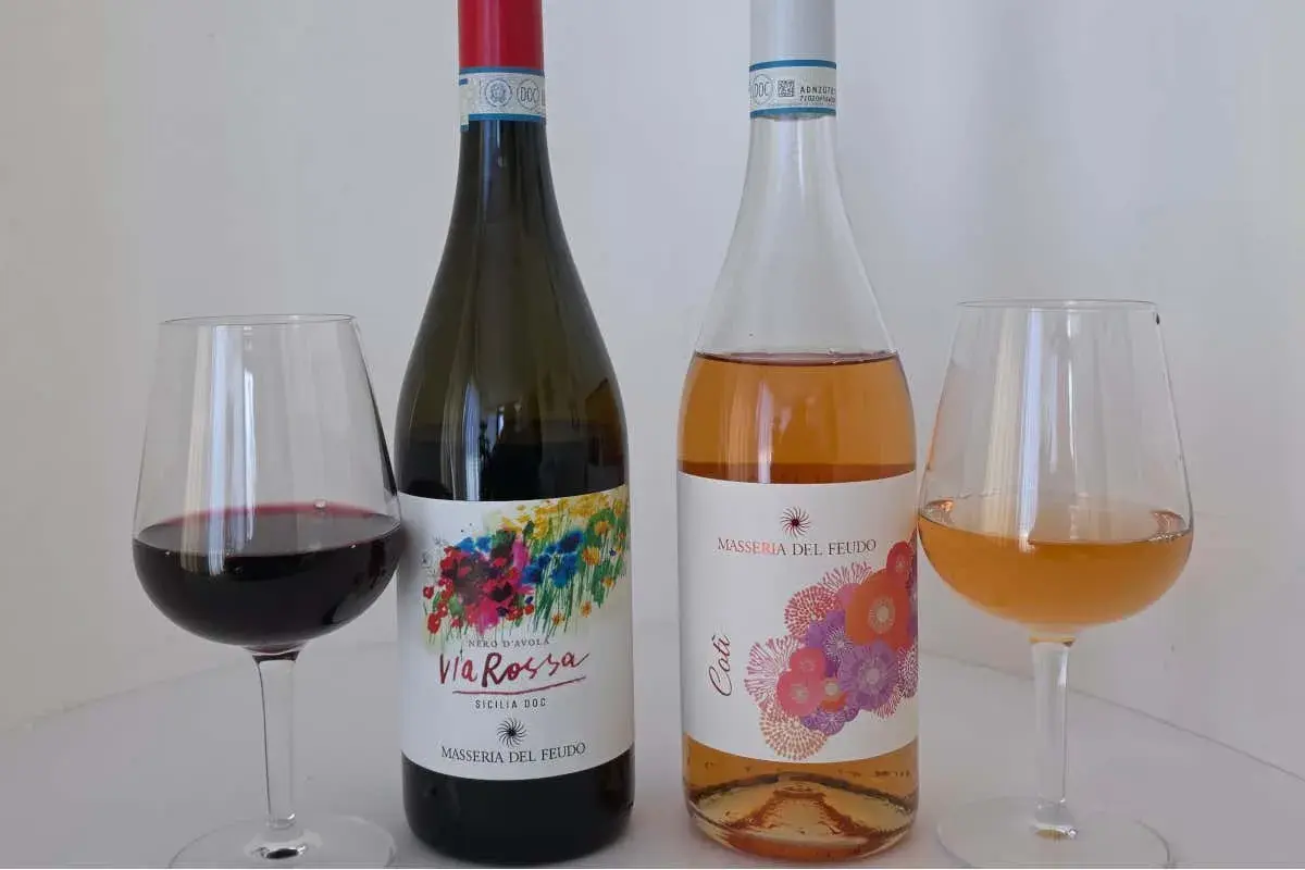 Due bottiglie di vino, una rossa e una rosata, con i loro calici. La **Masseria del Feudo** presenta le sue etichette colorate.