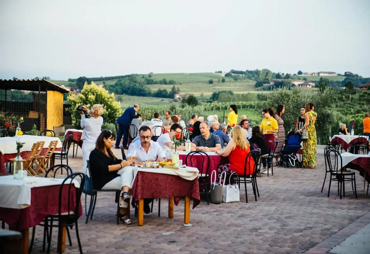 Un conviviale pranzo in vigna Monferrato, con tavoli apparecchiati all'aperto e persone che chiacchierano e gustano il cibo, sullo sfondo dolci colline.