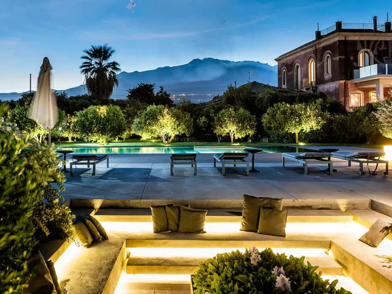 Un incantevole wine resort in Sicilia, con piscina illuminata, lettini e una vista mozzafiato sul vulcano al tramonto.