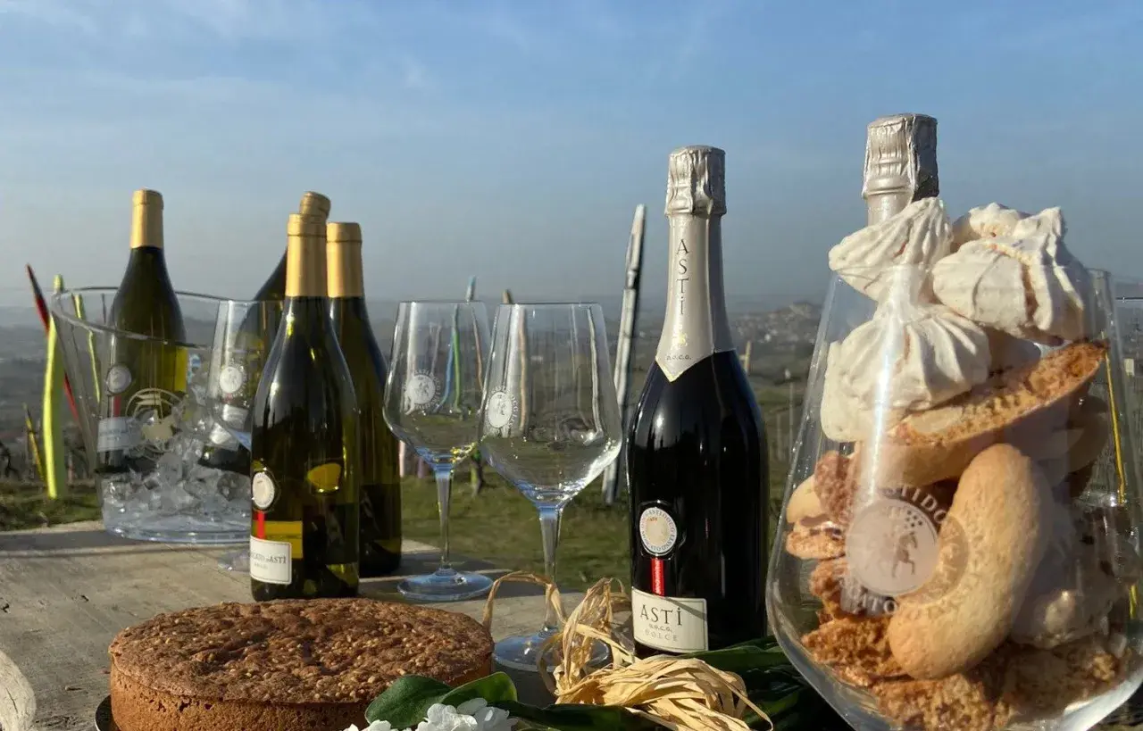Bottiglie di vino dolce Asti, bicchieri, torta e dolcetti in un allestimento all'aperto.
