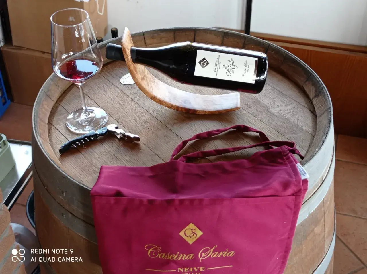 Bicchiere di vino rosso, cavatappi e borsa bordeaux con logo "Cascina Saria" su una botte di legno.