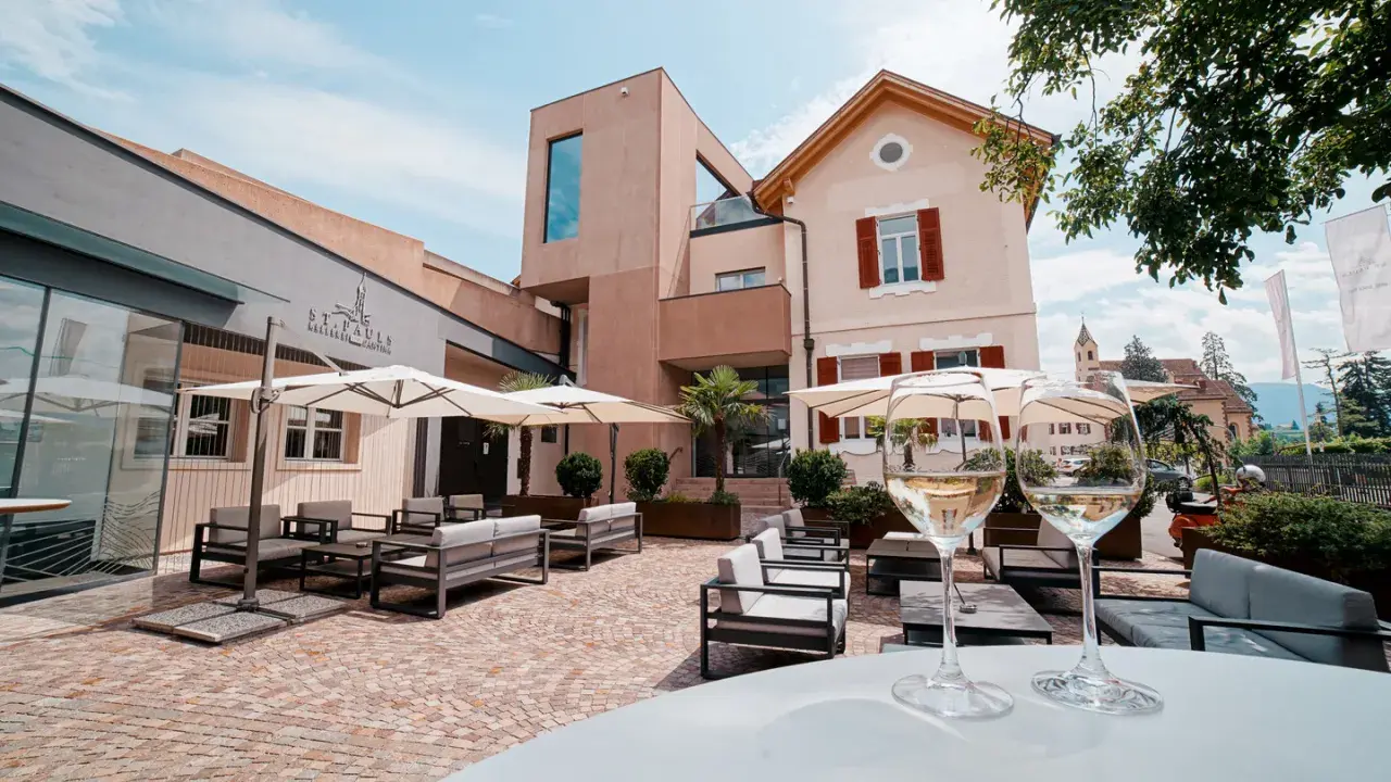 Terrazza accogliente con divani e ombrelloni, due calici di vino bianco in primo piano. La cantina St. Pauls sullo sfondo.