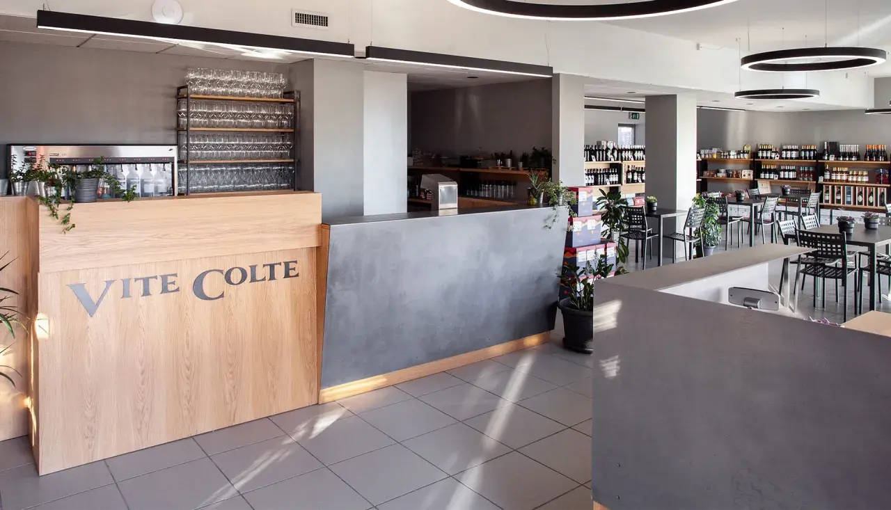 Interno di un negozio di vini con bancone in legno e cemento, scaffali pieni di bottiglie e tavoli pronti per accogliere i clienti. Il logo "Vite Colte" è ben visibile.