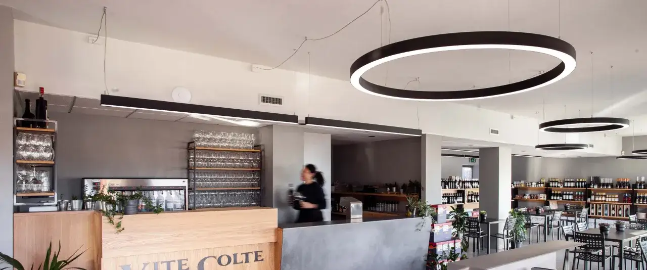 Interno di un locale moderno con bancone in legno e cemento, scaffali di bicchieri e bottiglie. Si intravede una persona che serve, in un ambiente che evoca la degustazione di vite colte.