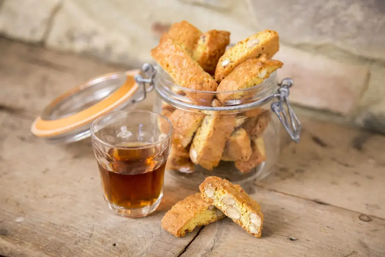 Cantucci e vino, un classico intramontabile. Biscotti croccanti con mandorle in un barattolo di vetro, accanto a un bicchierino di vino ambrato.