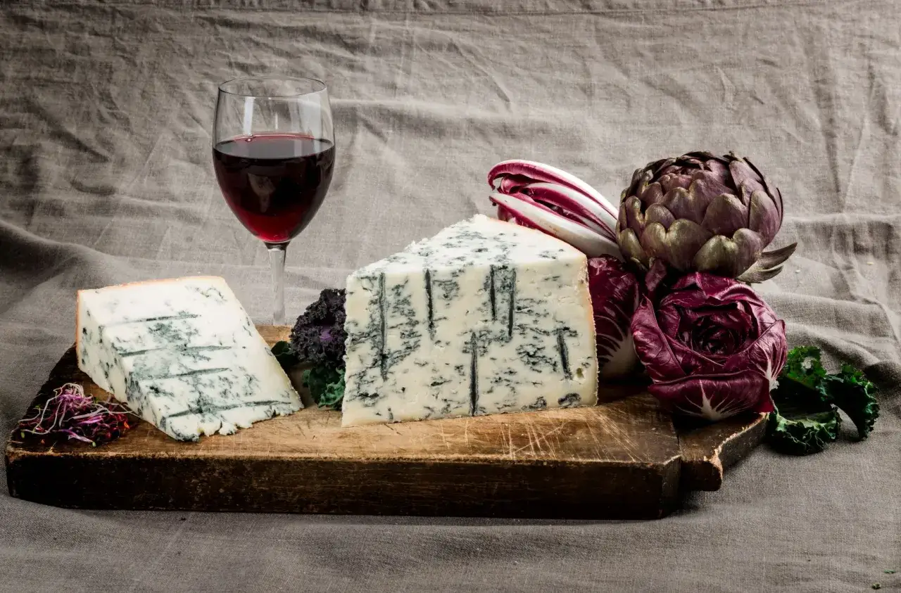Fetta di gorgonzola su tagliere di legno, con bicchiere di vino rosso e verdure. Un perfetto gorgonzola abbinamento vino.