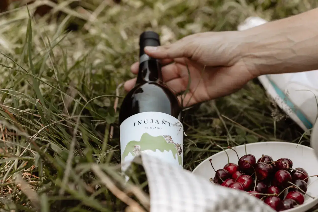 Un picnic sull'erba con una bottiglia di vino Friulano e una ciotola di ciliegie. Il profumo del vino e della frutta evoca un fragrante significato di piacere estivo.