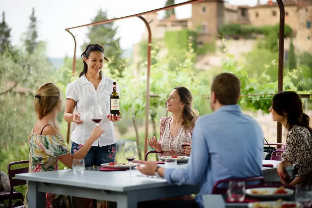 Degustazione di vino in una delle cantine di Greve in Chianti. Un gruppo di amici sorride mentre una cameriera presenta una bottiglia di Chianti Classico.