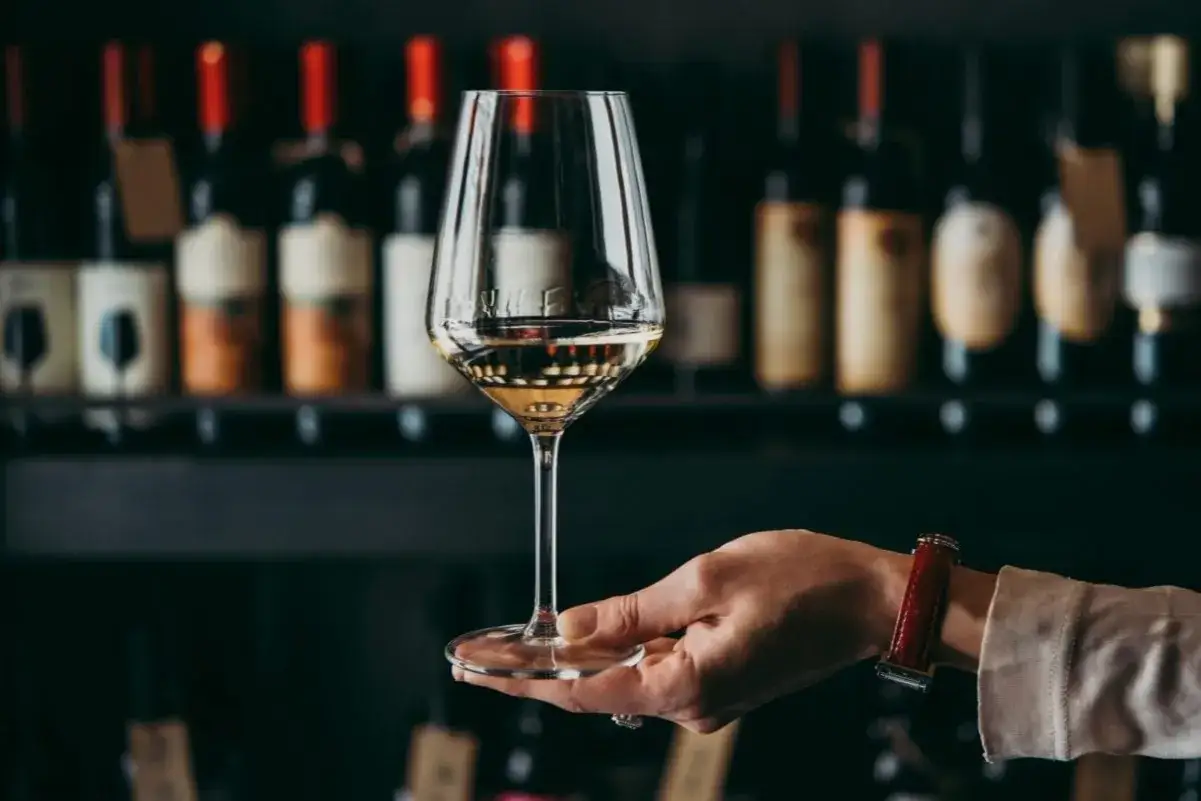 Un bicchiere di vino bianco dorato, tenuto da una mano con un orologio rosso, sullo sfondo di bottiglie di vino.