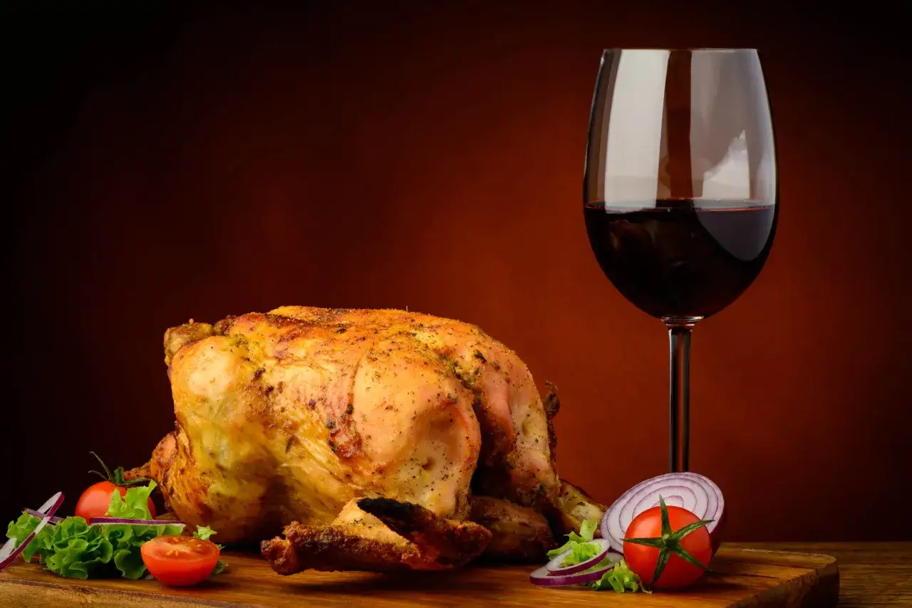 Pollo arrosto dorato su tagliere con contorno di verdure fresche e un bicchiere di vino rosso, perfetto vino da abbinare al pollo.