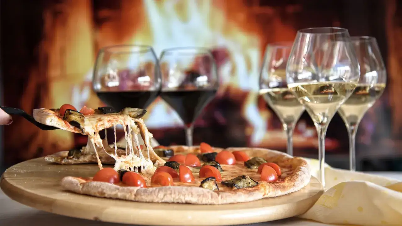 Una fetta di pizza filante con melanzane e pomodorini, accompagnata da calici di vino, perfetta per una serata davanti al camino.